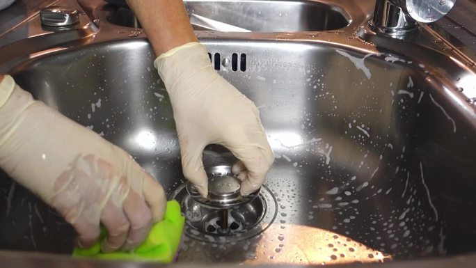 一个女人戴着手套的手在厨房用海绵和清洁剂清洗金属水槽的特写镜头。缓慢的运动。