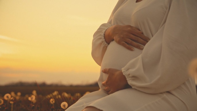 身穿白色连衣裙的孕妇在朝阳的田野里揉着肚子