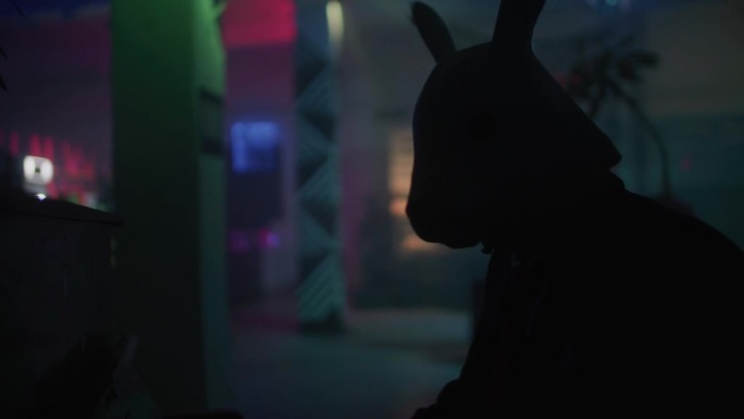 一名穿着西装、戴着兔子面具的男子在俱乐部里弹钢琴，看着镜头