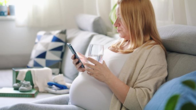 孕妇在沙发上喝水和使用智能手机