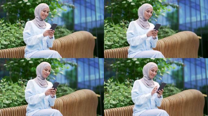 戴着头巾的年轻穆斯林女职员坐在办公大楼附近的长椅上，微笑着使用智能手机