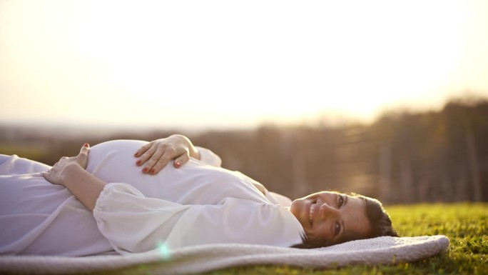 悠闲的孕妇穿着白色连衣裙躺在阳光明媚的田野里