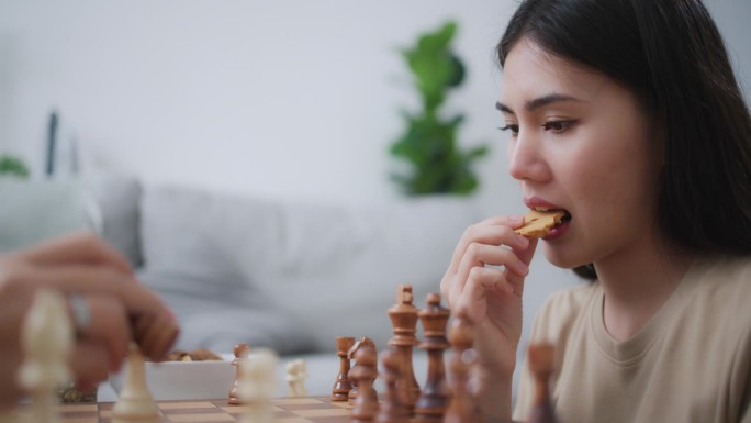 年轻的亚洲女子在家里和朋友下棋时吃着零食