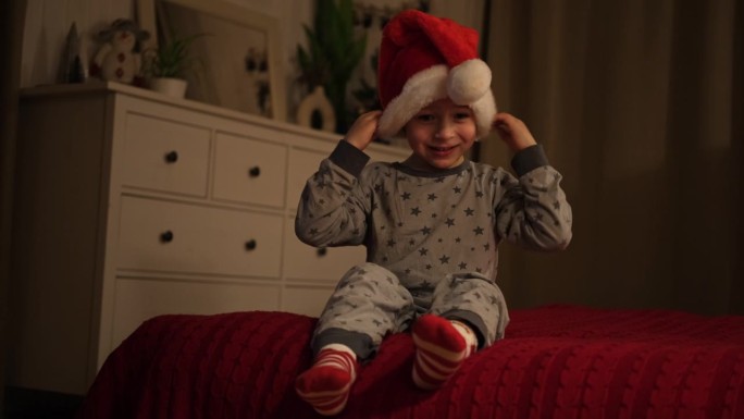 圣诞快乐。滑稽的小男孩边笑边拿着圣诞礼盒，玩得很开心。新年快乐。兴奋的小孩穿着睡衣，在床上跳来跳去。