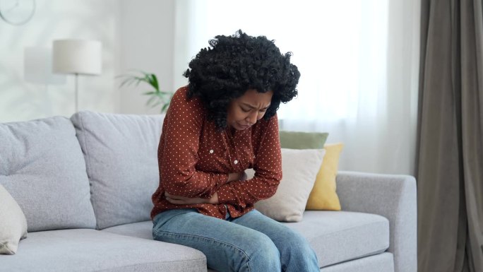 一位年轻的非裔美国女性，因腹痛而坐在家里房间的沙发上。