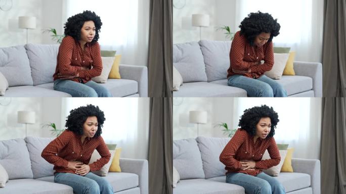 一位年轻的非裔美国女性，因腹痛而坐在家里房间的沙发上。