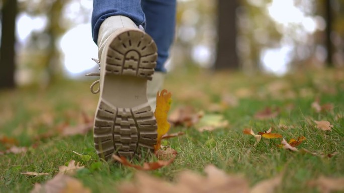 在草地上的秋天公园里，现场摄像机跟随一个穿着白色靴子的女孩的脚慢动作行走。后视图跟踪拍摄的孩子漫步在