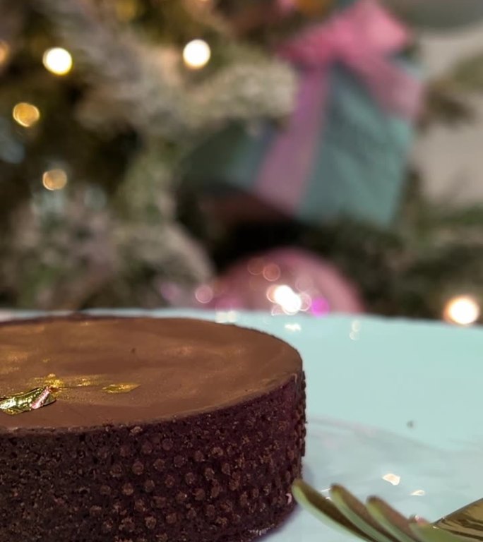 巧克力蛋糕放在蓝色的盘子上，背景是一棵新年圣诞树闪烁的彩灯广告，金色的叉子反射的光在蛋糕上烘烤着美味