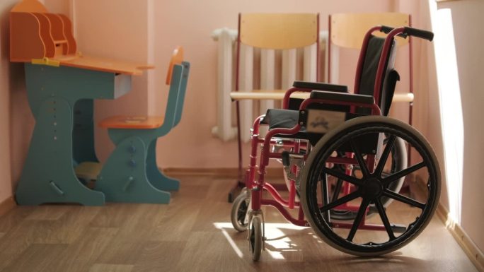 一辆儿童轮椅矗立在学校走廊的中央，背景是一张课桌。儿童康复中心。