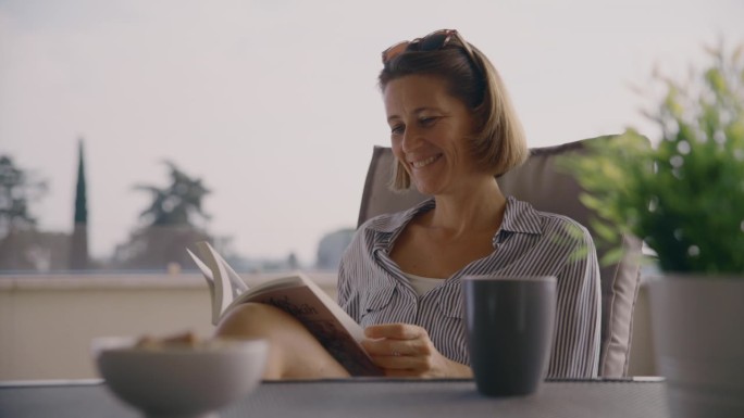 快乐的中年成年女性在家里的露台上一边喝咖啡一边看书
