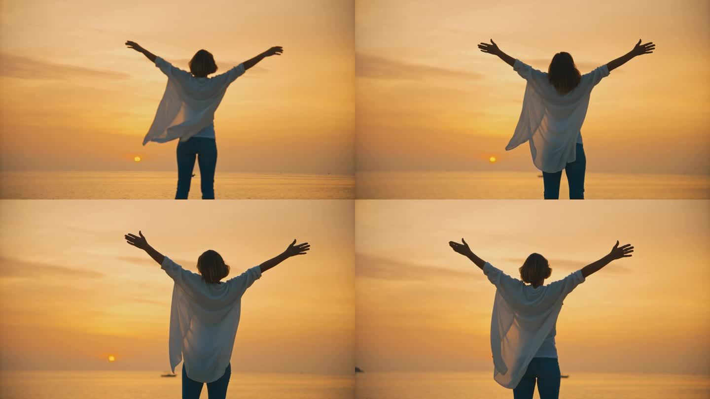 在克罗地亚罗维尼的夕阳下，一名无忧无虑的女游客站在海边，张开双臂，面对着广阔的橙色天空