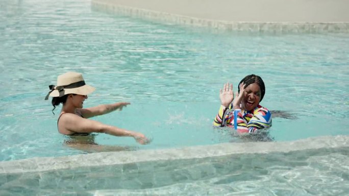 西班牙裔女子站在池水里，用手舀起水泼向站在前面的非洲裔美国朋友，非洲裔女子无法还击，只好笑着用手捂住