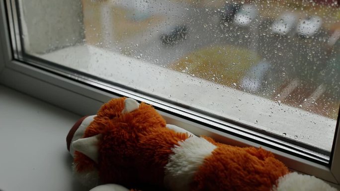 从房子内部到窗户到街道的视野。窗台上有一个柔软的玩具;秋天外面在下雨。雨滴落在玻璃上。温暖和舒适的房
