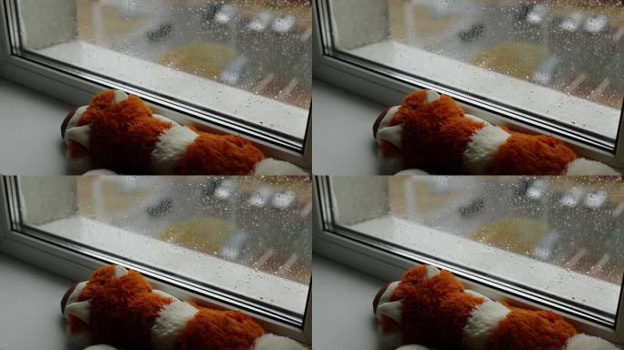 从房子内部到窗户到街道的视野。窗台上有一个柔软的玩具;秋天外面在下雨。雨滴落在玻璃上。温暖和舒适的房