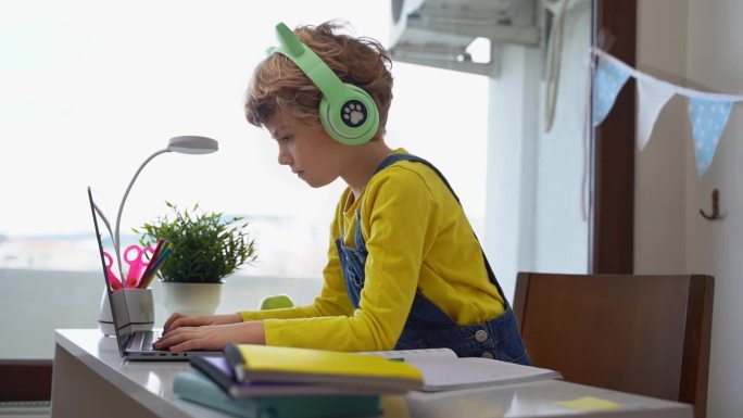 可爱聪明的白人小学生戴着耳机在笔记本电脑上打字。在线家教教授数字课，在家电脑上网络课。虚拟教育。学习
