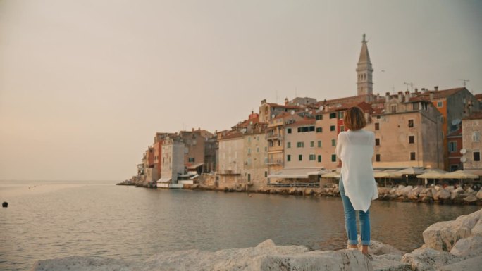 女游客站在克罗地亚海边的岩石上，平静地看着罗维尼的老城