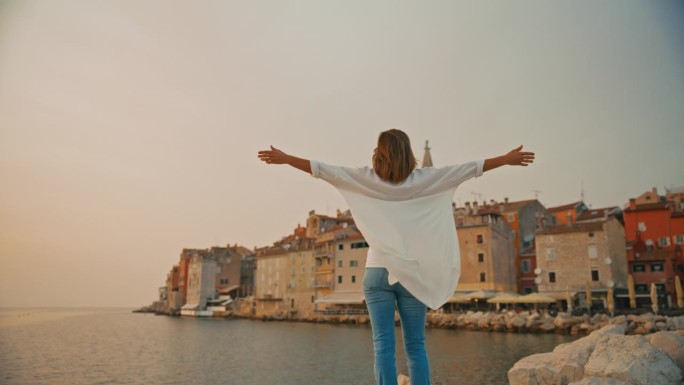 克罗地亚罗维尼，一名女游客站在老城区和晴朗的天空前伸展双臂