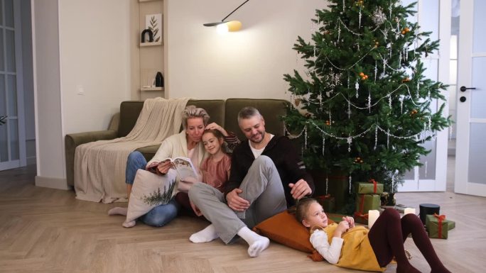 一个友好的家庭坐在圣诞树旁边的地板上看书。