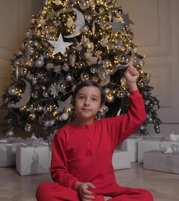 圣诞快乐，孩子。圣诞节那天，一个穿着红色睡衣的男孩坐在家里的圣诞树旁。他对着镜头挥手打招呼，手指向上