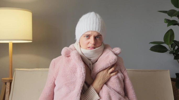 悲伤的女人穿着冬天的外套和帽子，在家里的沙发上感到寒冷，因为缺乏热量，看着相机试图获得温暖，表达负面