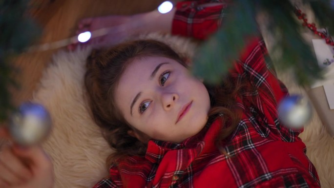 特写镜头。女孩躺在圣诞树旁，如梦似幻地玩着圣诞球。