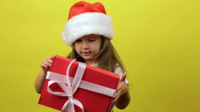 可爱的小女孩孩子戴着圣诞老人的帽子，拿着礼盒，在工作室的黄色背景墙上摆出孤立的姿势。圣诞节的概念