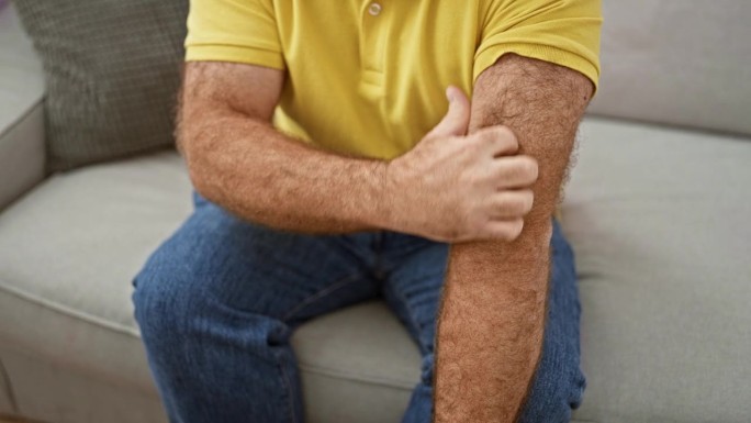 英俊的中年白人男子坐在家里的沙发上，抓挠手臂上发炎的皮肤。瘙痒性皮炎皮疹引起不适。过敏反应需要保健。
