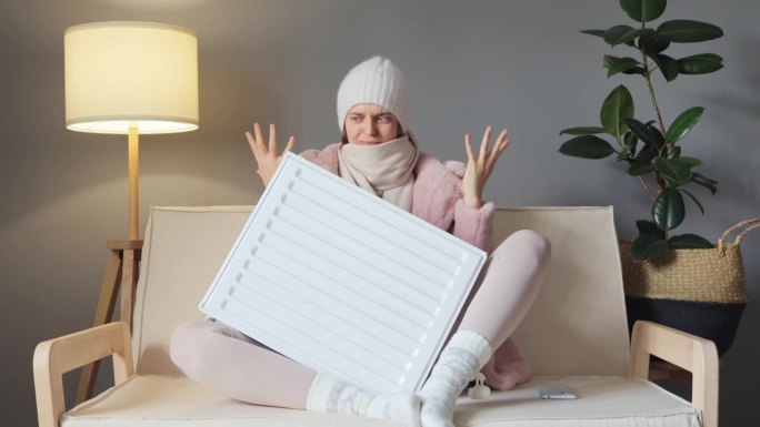 愤怒的女人穿着冬天的羊毛大衣，戴着帽子，坐在电油加热器前，看起来很伤心，被冻住了。用消极的面部表情惊
