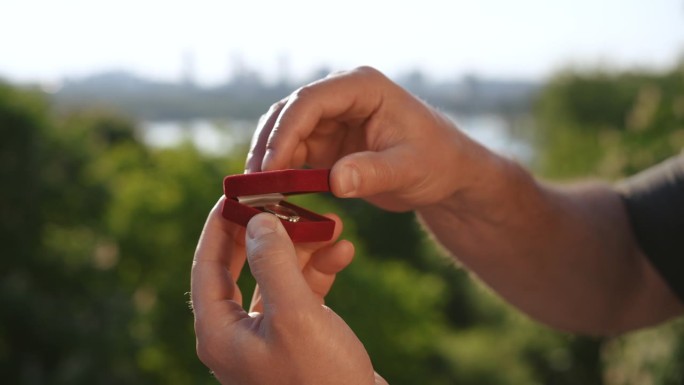 一个男人的手打开一个天鹅绒盒子的特写，里面有一个戒指，背景是河边的一个城市。