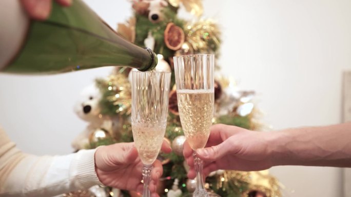 手拿着瓶子，把香槟酒倒进玻璃杯里，叮当作响的玻璃杯背景是一棵装饰过的圣诞树。用酒杯打人。碰杯祝酒时碰