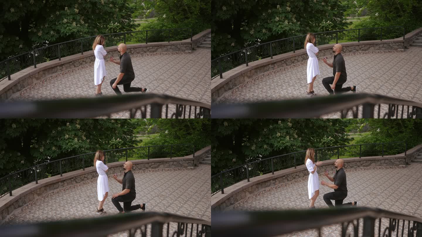 在慢镜头中，一个男人在城市公园单膝跪地向他的女人求婚，并给了他订婚戒指。爱情和婚姻的概念。