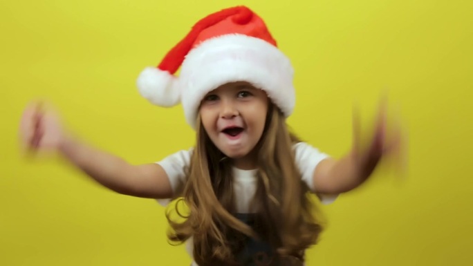 蹦蹦跳跳的小女孩戴着圣诞老人的帽子，在黄色的背景工作室里跳舞，玩得很开心，有表现力的手势。新年快乐的