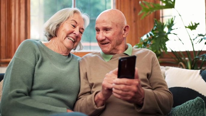 老年夫妇，在家里用智能手机聊天，在沙发上阅读通知和新闻应用。快乐的老人，女人，在客厅里用手机打字，社