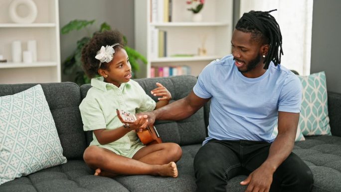 非裔美国父亲和女儿坐在沙发上教他们弹尤克里里