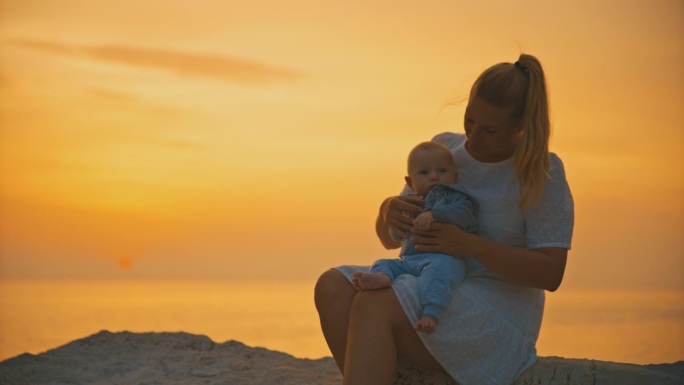 克罗地亚罗维尼，日落时分，慈爱的母亲带着刚出生的婴儿坐在海边的岩石上，映衬着橙色的天空