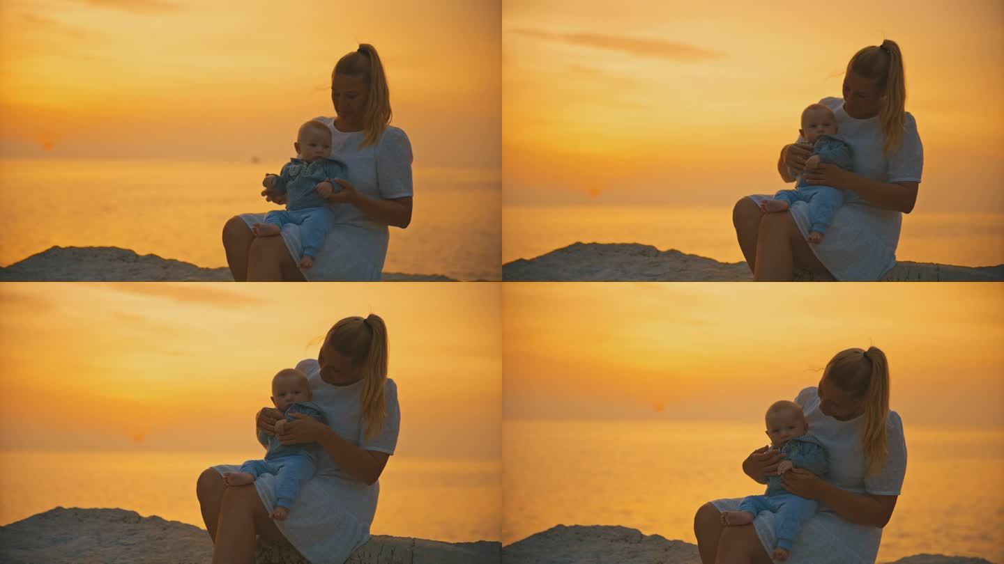 克罗地亚罗维尼，日落时分，慈爱的母亲带着刚出生的婴儿坐在海边的岩石上，映衬着橙色的天空