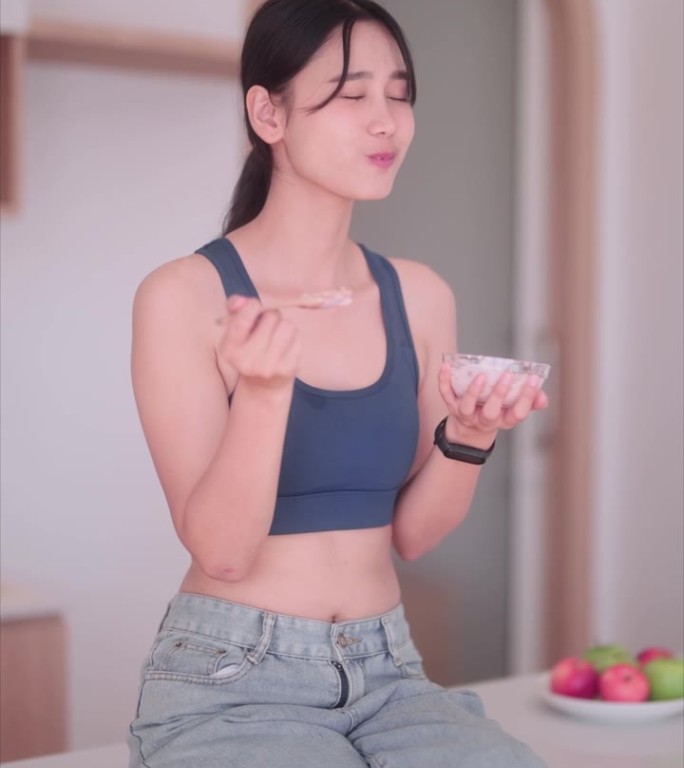 穿着运动服的亚洲女运动员在舒适的厨房里品尝着清爽的酸奶。她崇尚健康的生活方式，用有益健康的食物滋养自