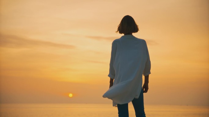克罗地亚罗维尼，日落时分，一名女游客站在海边，双手插在头发里，面对着广阔的橙色天空