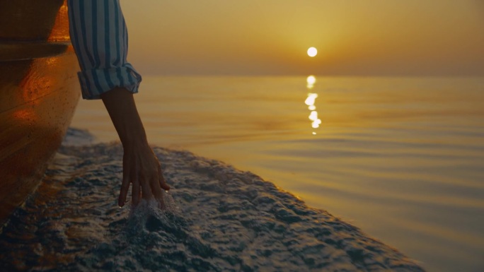 日落时分，一名无法辨认的女子在船上航行，用手触摸着海面