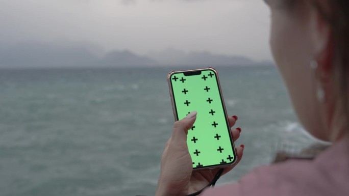 从一个女人的头后面看，她在智能手机上滚动着一些东西，绿色的屏幕是一个模型，背景是海边阴天的夜晚。高悬