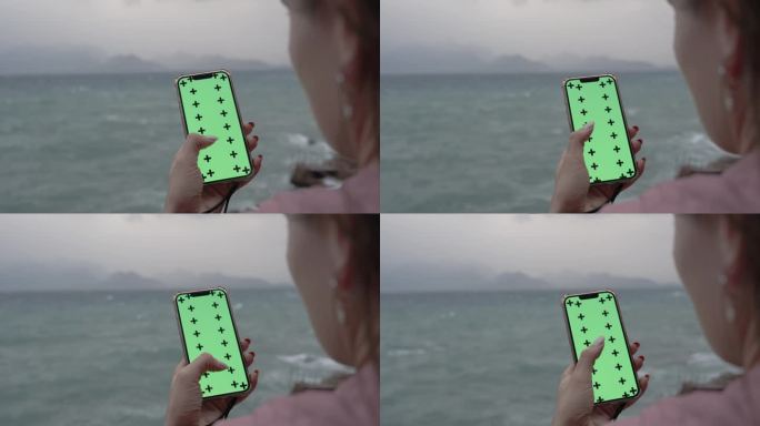 从一个女人的头后面看，她在智能手机上滚动着一些东西，绿色的屏幕是一个模型，背景是海边阴天的夜晚。高悬