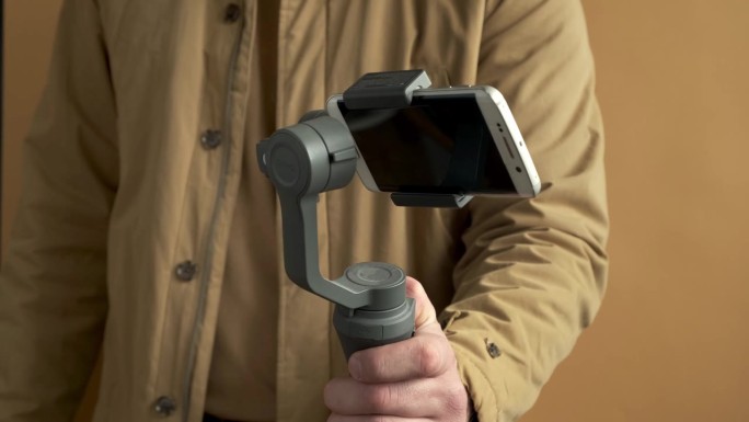 一名男子用智能手机在黄色背景上使用万向节稳定器进行拍摄。使用三轴电子稳定器进行vlog或视频拍摄。