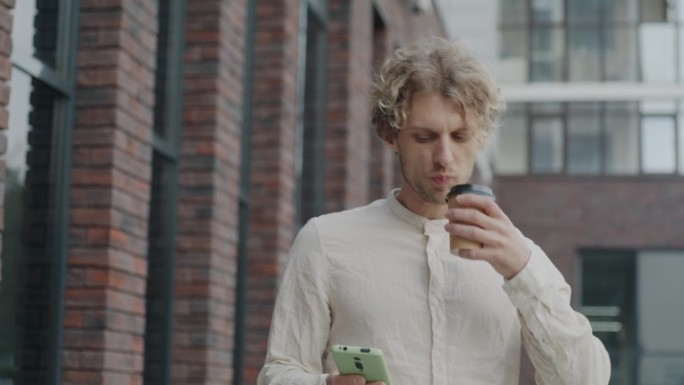 多莉拍摄的年轻商人走在城市街道上喝着带走的咖啡，用智能手机上网聊天