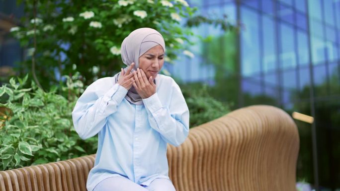 戴着头巾的年轻穆斯林女职员坐在办公楼附近的长椅上，牙疼得难受。