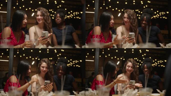 三个不同的女人晚上在餐厅见面，在智能手机上看照片，笑