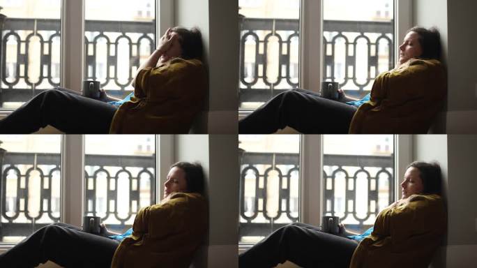 中年沮丧的妇女坐在靠近窗户的地板上，用手遮住脸，闭上眼睛，痛苦地忍受着绝望。