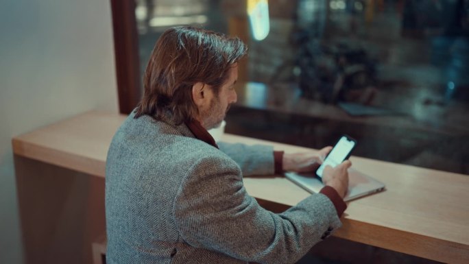 成熟商人在咖啡店透明窗户附近使用智能手机的后视图