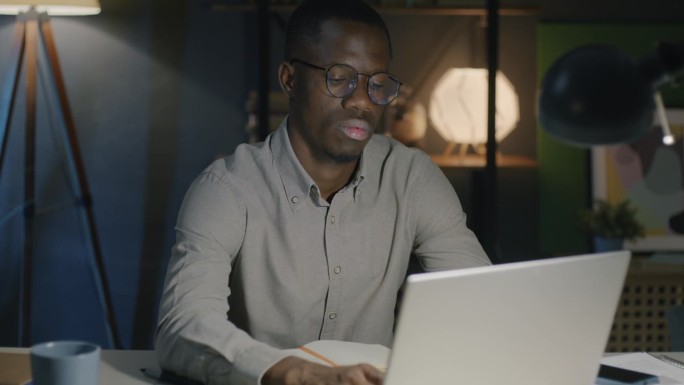 一位非洲裔美国商人，晚上在灯火通明的办公室里，用笔记本电脑打字，专注于网上工作