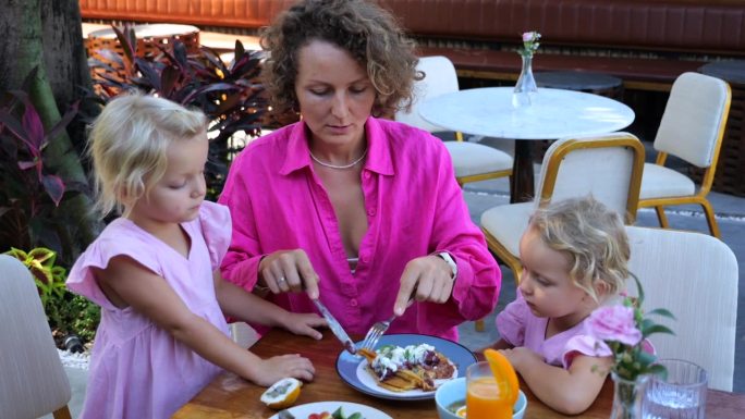 年轻的母亲切下一块美味的煎蛋卷，在咖啡馆喂女儿。在夏日咖啡馆喂孩子的动态视频。母亲和女儿在咖啡馆度假