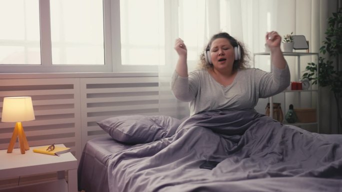 快乐的超重女人在手机应用上听着最喜欢的歌曲，在床上跳舞